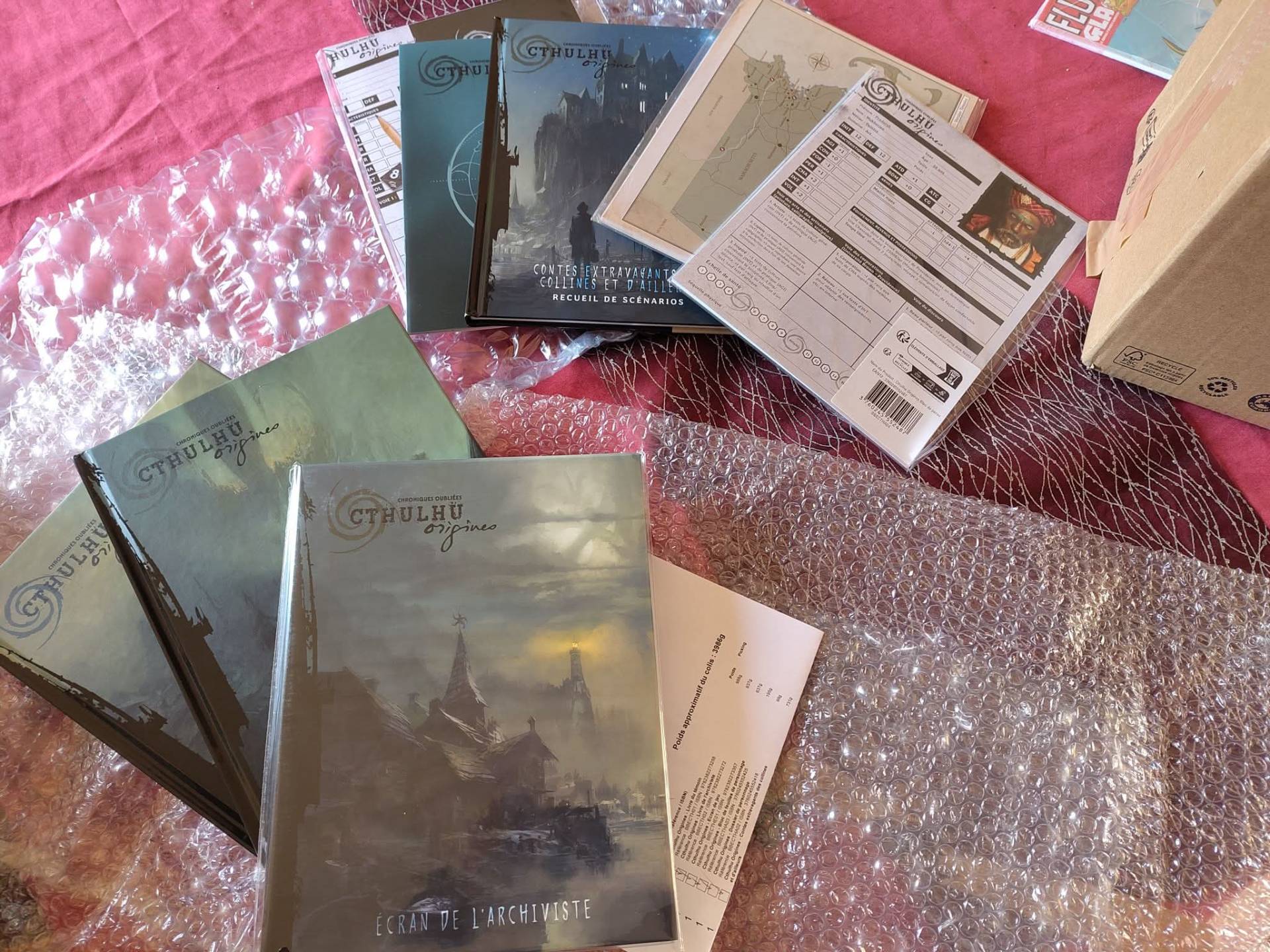 Manuels et écran de jeu de la gamme Cthulhu Origines, fraîchement déballés sur un tissu rouge